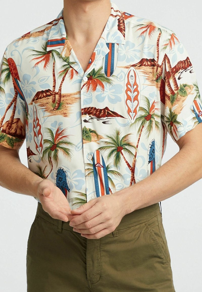 Homme portant une chemise boutonnée à manches courtes avec un imprimé de palmiers, planches de surf et volcans, associée à un pantalon vert olive.