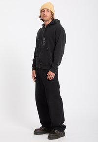 Jeune homme debout portant un bonnet beige, un sweat à capuche noir zippé avec des poches devant, un pantalon noir ample et des chaussures noires à lacets.