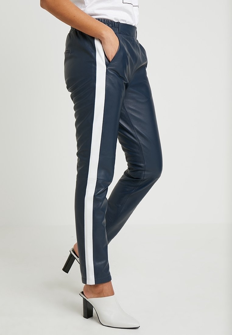Marineblauwe faux leren broek met witte zijstrepen; elastische tailleband; slim fit; voorzien van zijzakken; gedragen met witte hoge hakken.