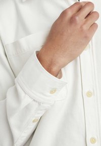 Chemise blanche à boutons avec une finition texturée, dotée d'une poche poitrine et de coutures discrètes. Les poignets ont des boutons jaunâtres.