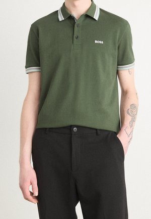 Mann trägt ein olivgrünes BOSS Polohemd mit weiß gestreiftem Kragen und Ärmelbündchen, schwarze Hose und sichtbares Schlangentattoo am rechten Unterarm.