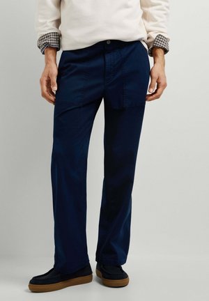 Mann trägt dunkelblaue gerade geschnittene Hose mit aufgesetzten Taschen und schwarze Schuhe mit braunen Sohlen, steht vor einem einfarbigen Hintergrund.
