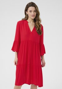 Robe rouge, longueur genou, en tissu léger. Présente un col en V, des manches volantes et un design à niveaux pour plus de texture.