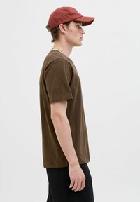 Bruine katoenen T-shirt met korte mouwen, relaxte pasvorm. Gecombineerd met een rode baseballcap. Model staat links, draagt zwarte broeken.