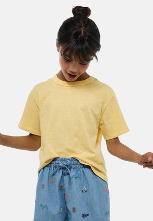 Enfant aux cheveux foncés en chignon portant un t-shirt jaune et un short bleu avec des broderies colorées sur le thème du camping, regardant vers le bas avec les bras légèrement levés.