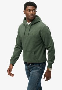 Felpa con cappuccio verde realizzata in tessuto texture, con tasca frontale e cordini. Abbinata a jeans blu, che mostrano un fit casual.