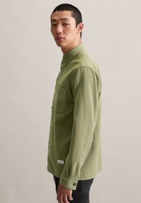Green textured shirt with a button-up design, single chest pocket, and black buttons, worn with dark jeans.