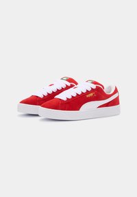 Red suede sneakers feature white laces, a textured rubber sole, and a white side stripe. Green and white accents on the tongue.
