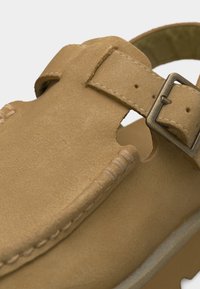 Λεπτομέρεια από καφέ σουέντ σανδάλι με ραμμένα άκρα και ρυθμιζόμενο λουρί με μεταλλική αγκράφα.