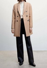 Abrigo beige de mezcla de lana con diseño de doble botonadura, solapas con muesca, botones negros y bolsillos laterales, combinado con pantalones anchos de cuero negro.