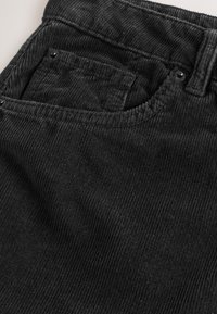 Gros plan sur la poche d’un pantalon en velours côtelé noir avec passants de ceinture visibles, rivets et détail du tissu texturé.