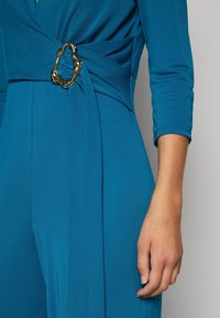 Robe portefeuille en teal en tissu lisse avec un détail de taille torsadée, mettant en avant une boucle ovale dorée métallique.