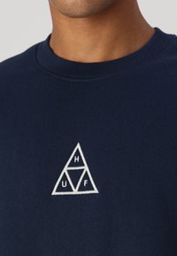 Sudadera azul marino de algodón, con un logo triangular bordado en blanco que contiene las letras H, U y F en su interior. Diseño de cuello redondo.