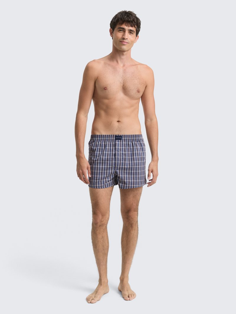 Jonge man die blootsvoets staat, gekleed in marineblauwe en witte geruite boxershorts tegen een effen lichte achtergrond.
