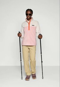 Pullover in pile bianco e rosa chiaro con colletto arancione, pantaloni beigi e scarpe da trekking marroni; tiene dei bastoni da trekking e indossa occhiali da sole.