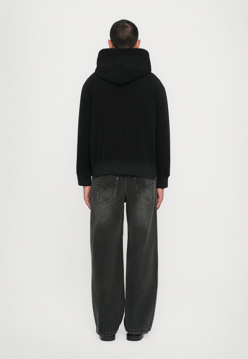 Schwarzer Hoodie mit großer Gesäßtasche und gerippten Bündchen, kombiniert mit lockeren grauen Jeans, die über Gesäßtaschen und Nahtdetails verfügen.