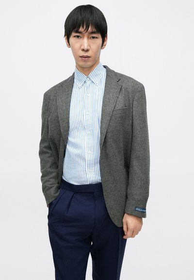 Blazer de lã cinzento sobre uma camisa azul claro com riscas brancas, com calças azul-marinho e uma etiqueta azul na manga do blazer.