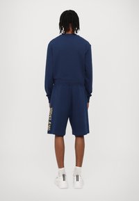 Temno modra majica in kratke hlače iz mehkega materiala, ki imajo zlat logo 'Versace Jeans' na levi strani hlač. Bele teniske.
