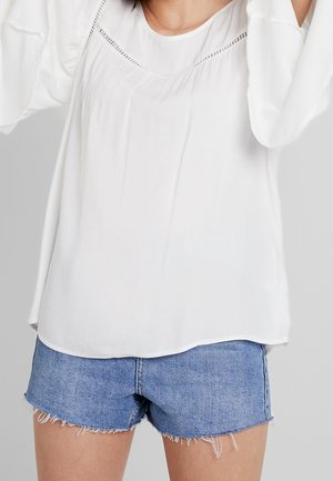 Kvinde iført en løs hvid bluse med lange ærmer og lyseblå slidte denimshorts mod en ensfarvet grå baggrund.