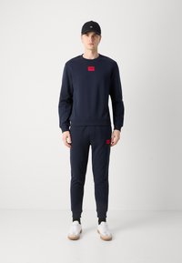 Completo felpa e joggers blu navy, in cotone, con un'etichetta rossa. Sneakers bianche completano l'outfit.