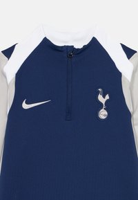 Mörkblå sporttopp med vita och grå ärmar, dragkedja framtill, Nike-swooshlogotyp på vänster sida och Tottenham Hotspur-emblem på höger bröst.