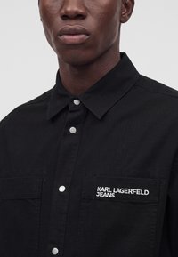 Близък план на човек, носещ черна риза с копчета и яка, с избродирано "KARL LAGERFELD JEANS" на джоба на гърдите.