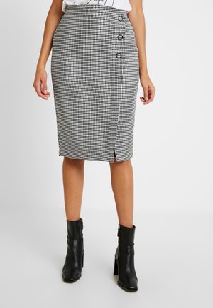 Jupe crayon en damier noir et blanc avec une fente sur le côté et trois boutons décoratifs à la taille, portée avec des bottines noires.