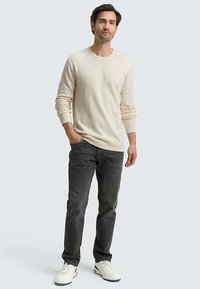 Suéter beige claro de cuello redondo con mangas largas, combinado con jeans gris oscuro y zapatillas blancas, posando frente a un fondo liso.