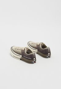 Zapatillas de deporte bajas con parte superior de lona beige, acentos marrones texturizados, detalle de franjas negras y marca en el talón. Suela de goma.