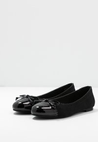 Ballerines noires avec un bout en verni brillant et un dessus en cuir suédé. Comprend un ornement en forme de nœud et des coutures décoratives le long des bords.