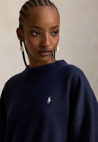 Μπλε σκούρο βαμβακερό φούτερ με φαρδιά λαιμόκοψη, ριμπ μανσέτες και μικρό κεντημένο λευκό λογότυπο στο στήθος. Υφή επιφάνειας και άνετη εφαρμογή.