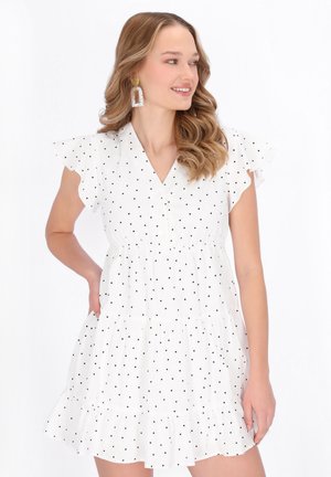 Naisella on aaltoilevat hiukset ja valkoinen lyhythihainen mekko, jossa on mustia pilkkuja ja röyhelöhalkio. Hän hymyilee ja katsoo oikealle.