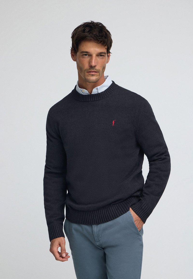 Dunkelgrauer Strickpullover mit Rundhalsausschnitt, gerippten Bündchen und Saum, mit einem kleinen roten Logo auf der Brust. Über einem hellblauen Hemd mit Kragen getragen.