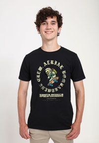 Černé bavlněné tričko s grafikou kostlivce ve skafandru na skateboardu, s textem v žluté a bílé. Kulatý výstřih a krátké rukávy.