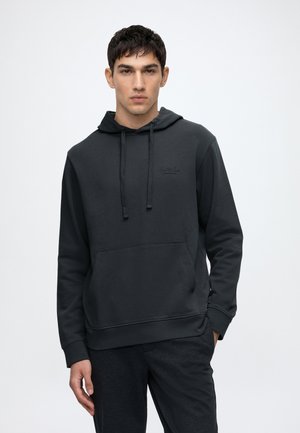 Jeune homme portant un sweat à capuche noir avec poche avant et cordons, debout avec une main dans la poche sur un fond clair uni.