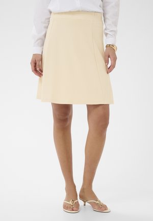 Ženska, ki nosi kremasto krilo dolžine do kolen, belo srajco z dolgimi rokavi, zlat zapestni trak in bež sandale z odprtimi prsti in peto.