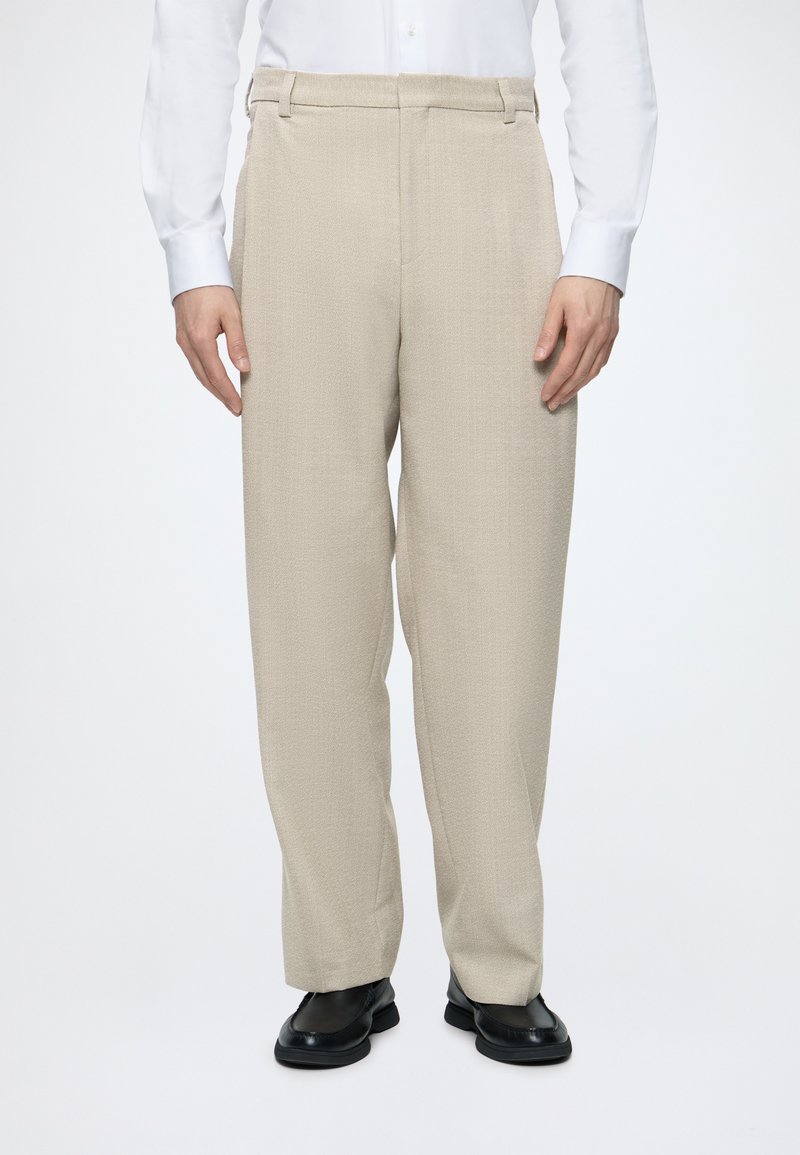 Homme portant un pantalon beige texturé, une chemise blanche et des chaussures en cuir noires, debout devant un fond blanc uni.