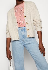 Cremefarvet ribbet cardigan med fem store knapper, lagdelt over en lyserød blomstret top, parret med lyseblå højtaljede jeans. Blød tekstur.