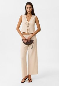 Beige Cropped-Top mit Schnürung und strukturiertem Gewebe, kombiniert mit weit geschnittenen Hosen und einer runden, geflochtenen Clutch-Tasche. Schwarze Sandalen mit Riemchen.