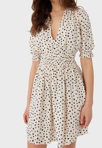 Cremfarbenes Kleid mit schwarzen Punkten, ausgestattet mit einem tiefen V-Ausschnitt, kurzen Puffärmeln und einem gerafften Bund. Der Stoff wirkt leicht und strukturiert.