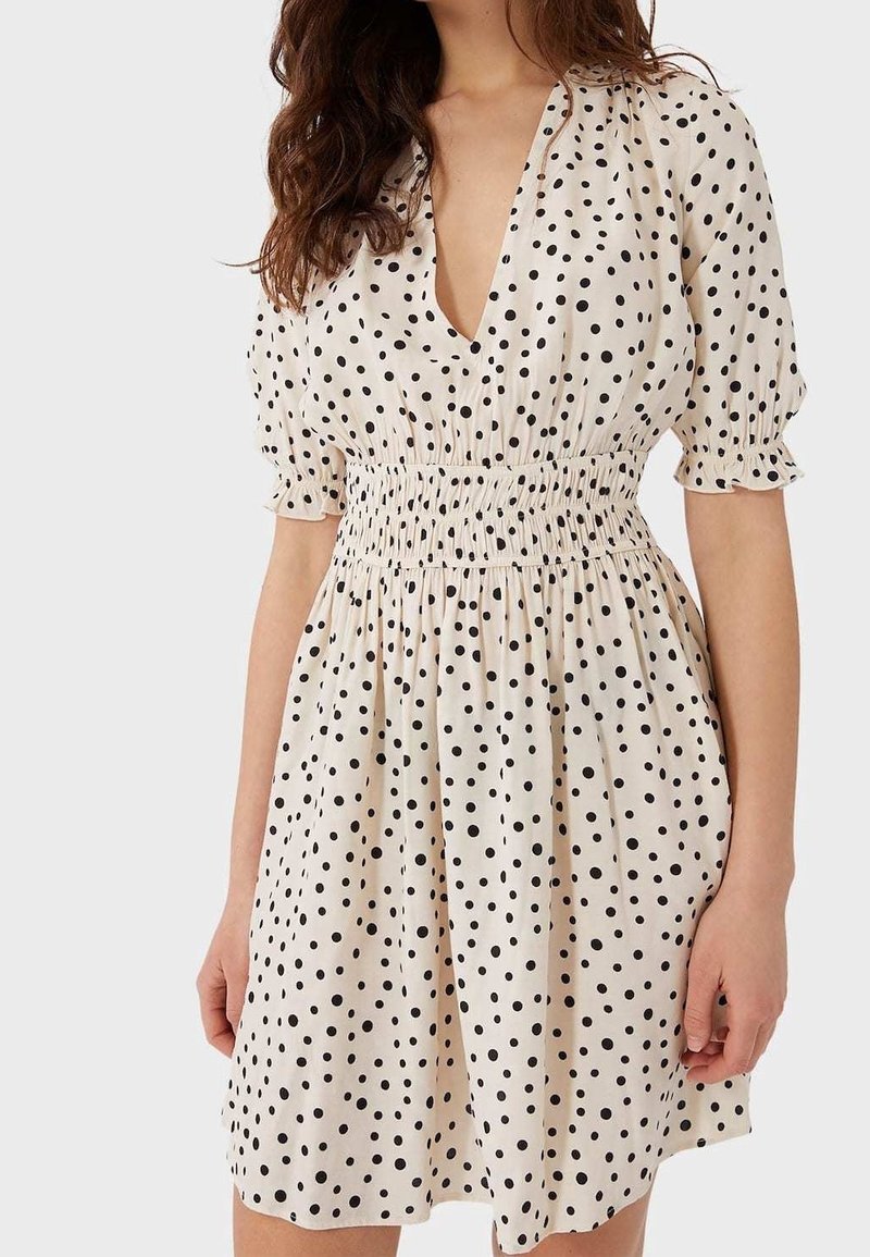 Cremfarbenes Kleid mit schwarzen Punkten, ausgestattet mit einem tiefen V-Ausschnitt, kurzen Puffärmeln und einem gerafften Bund. Der Stoff wirkt leicht und strukturiert.