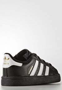 Svart Adidas Superstar-sneaker med vita ränder, guldlogotyp på hälen och "SUPERSTAR"-text på sidan, uppvisad mot en neutral bakgrund.