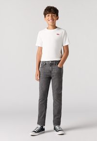 Camiseta de algodón blanca con logo rojo, combinada con jeans de mezclilla grises. Zapatillas altas en blanco y negro. Fondo liso.