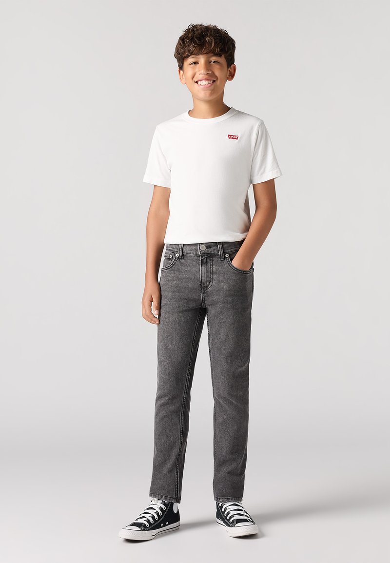 Camiseta de algodón blanca con logo rojo, combinada con jeans de mezclilla grises. Zapatillas altas en blanco y negro. Fondo liso.