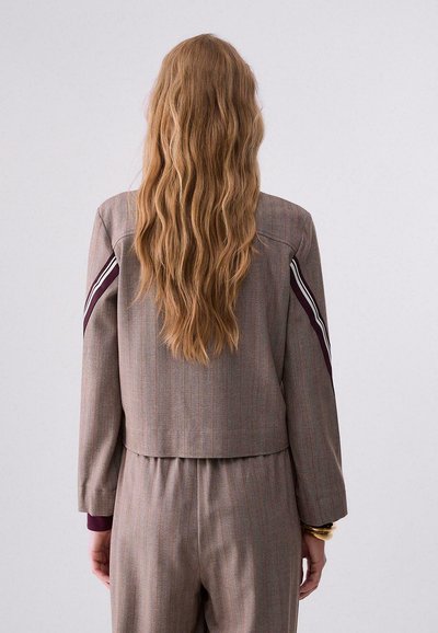 Personne aux longs cheveux ondulés châtain clair portant un costume gris à fines rayures avec des rayures bordeaux et blanches sur les manches, debout de dos.
