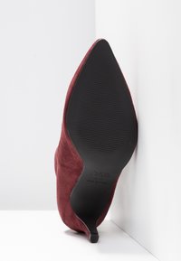 Chaussure à talon haut en daim bordeaux à bout pointu, inclinée contre des murs blancs, montrant la semelle noire avec les détails de la marque et de la taille.