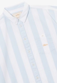 Chemise à rayures verticales bleu clair et blanches, à boutons, avec col blanc et poche poitrine brodée du texte "Blkvis" en orange.