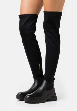 Bottes noires montantes au-dessus du genou avec semelles épaisses et fermetures éclair latérales, portées sur des jambes nues sur un fond clair uni.