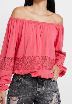 Kvinde iført en koralfarvet off-shoulder bluse med blonde detaljer og sorte slidte denimjeans, med hånden hvilende i lommen.