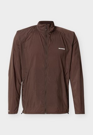 Giacca antivento leggera marrone con cerniera completa frontale, collo alto, tasche laterali con cerniera e logo Salomon bianco sul petto e sulla manica.